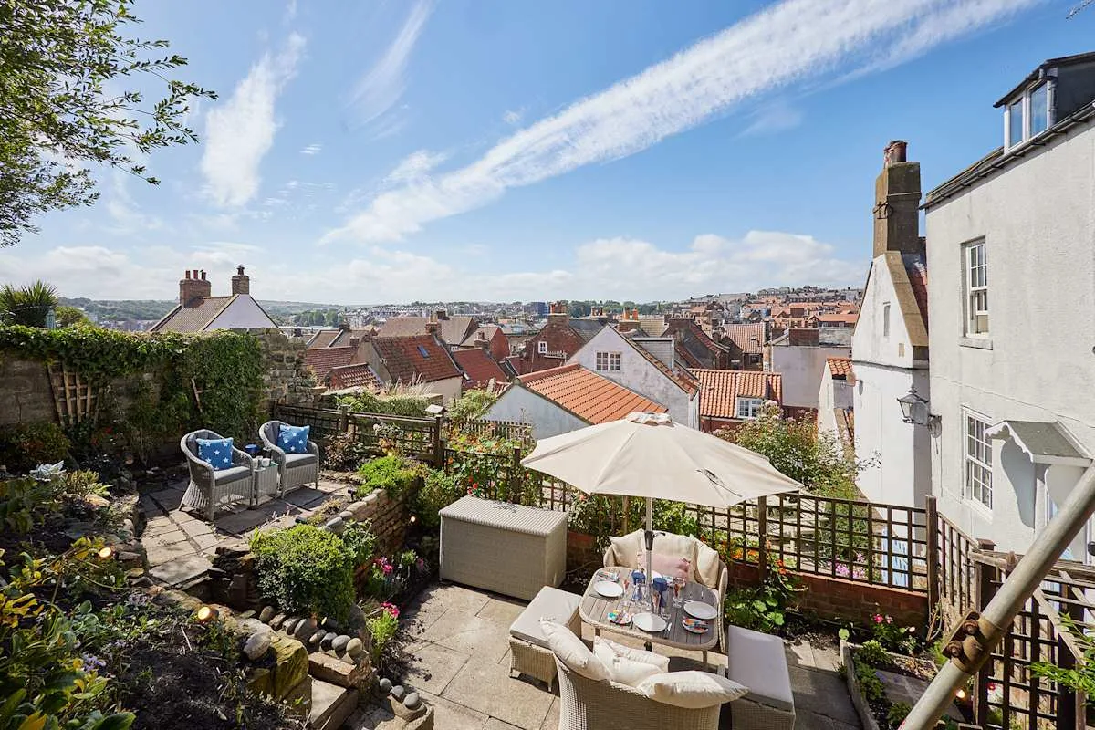 A garden terrace overlooks a town, with outdoor seating and a table under an umbrella.