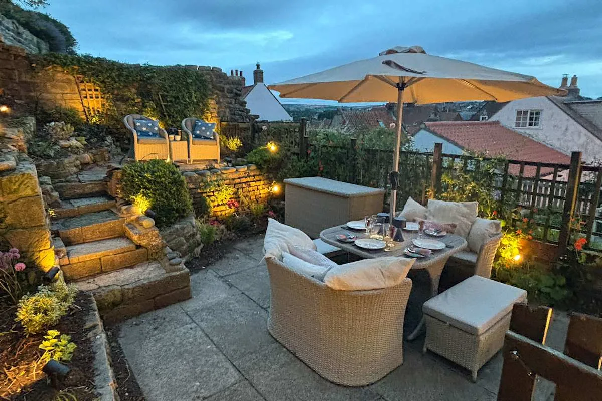 An outdoor patio has a dining table beneath a large umbrella, with seating and garden steps.