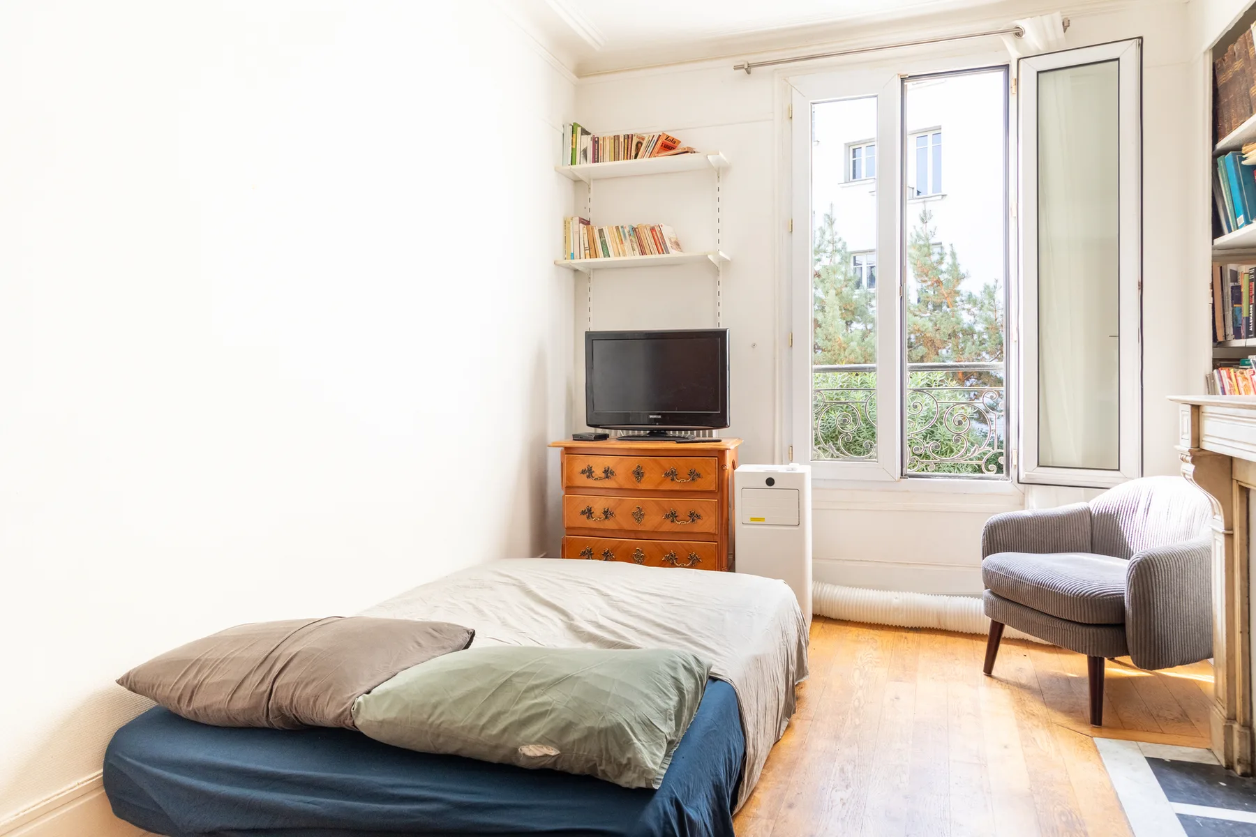 A bright bedroom with a bed, a television, and a chair beside tall windows.