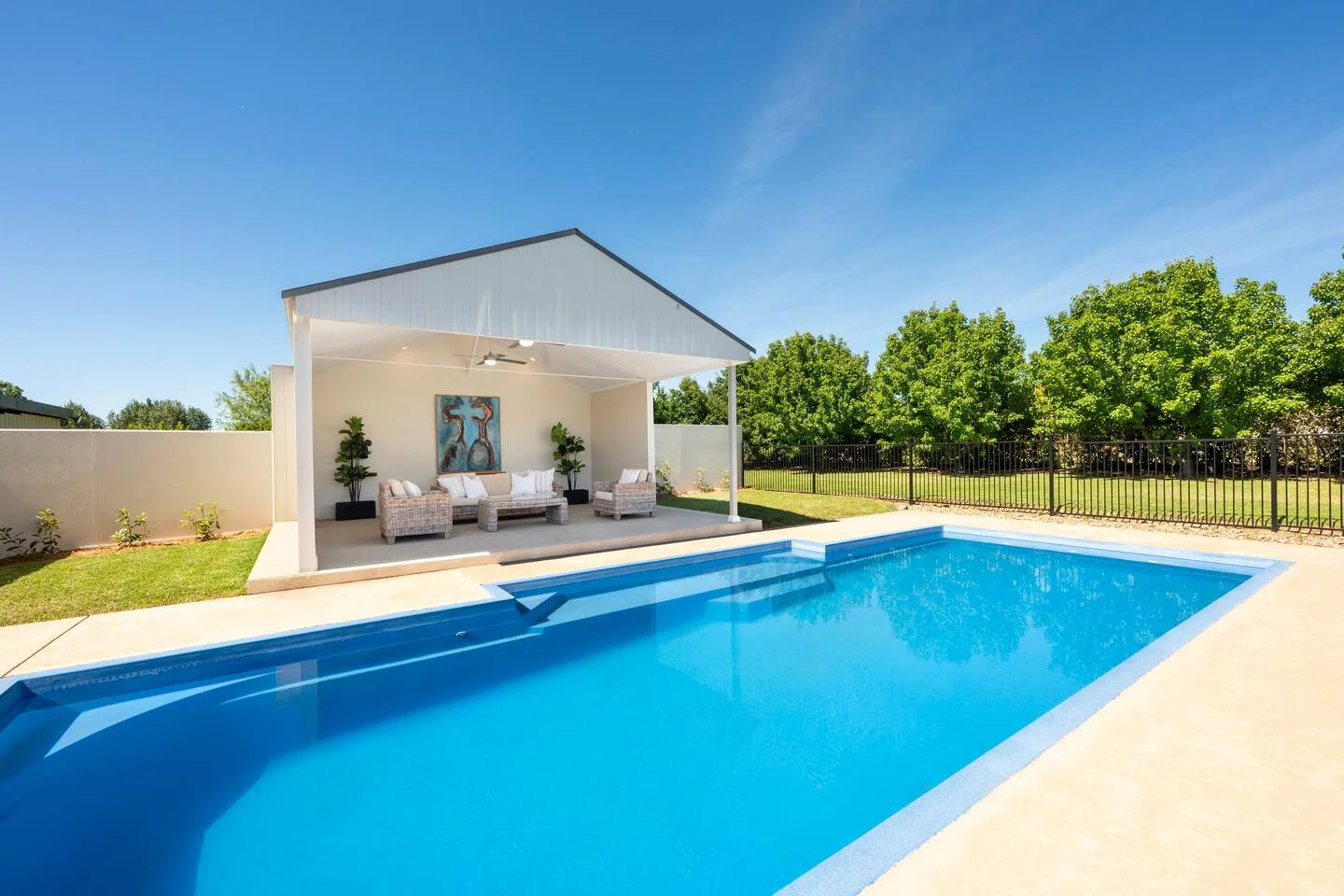 An outdoor swimming pool sits beside a covered seating area and lawn.