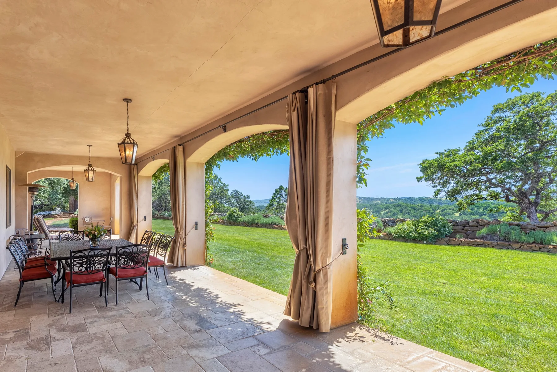 A covered patio with dining seating overlooks a green lawn and trees.