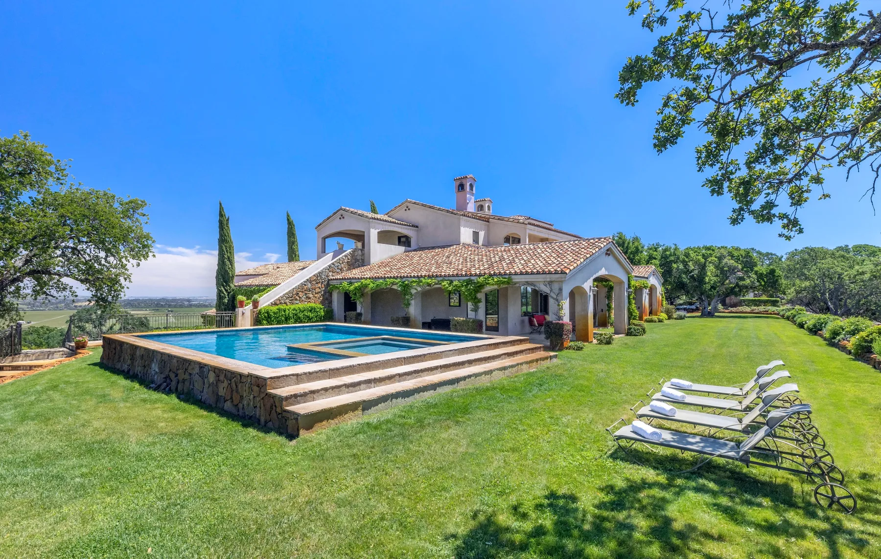A landscaped outdoor pool beside a large house, with lounge seating on the lawn.