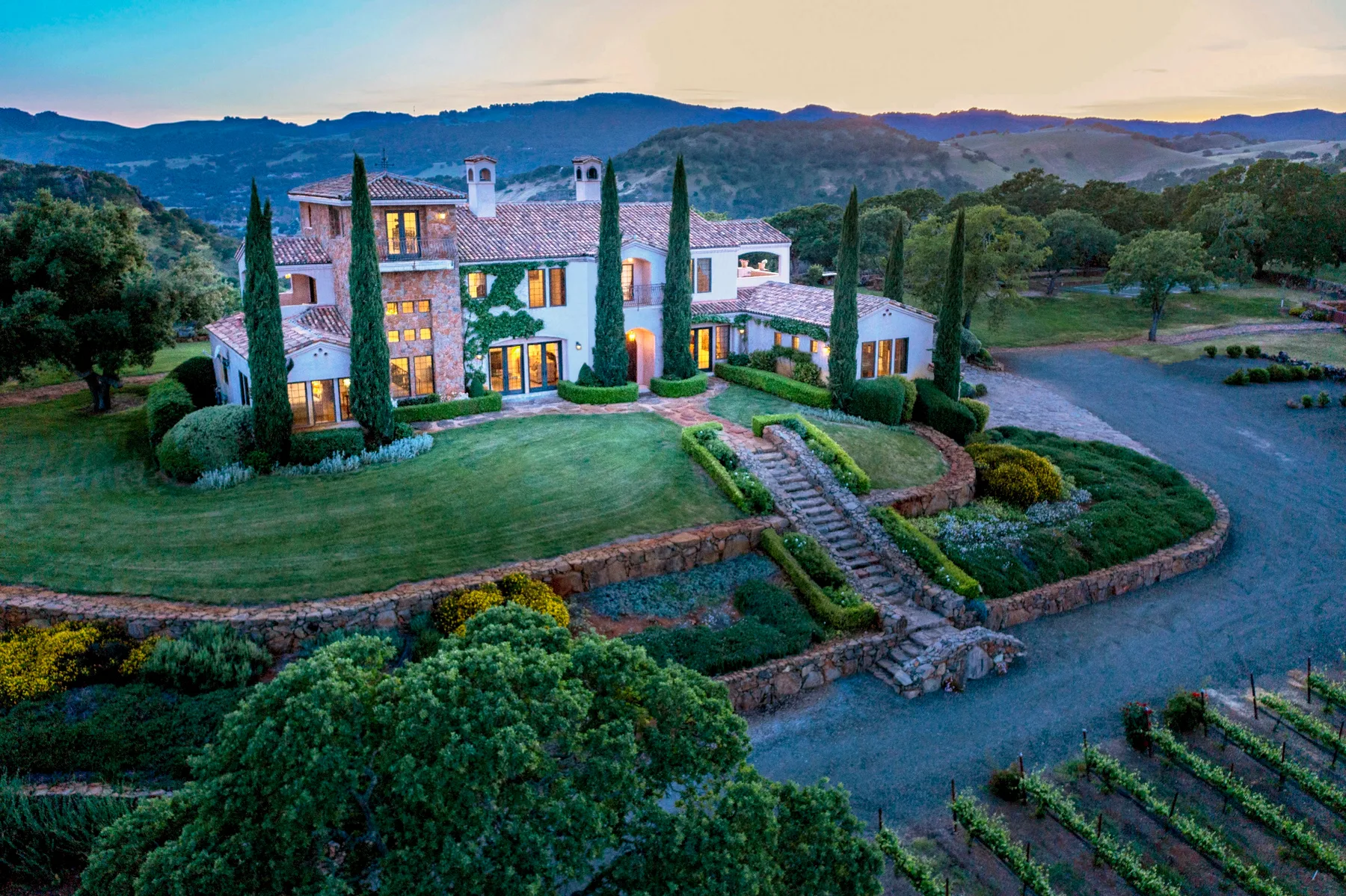 A large estate house stands amid landscaped gardens and rolling hills.