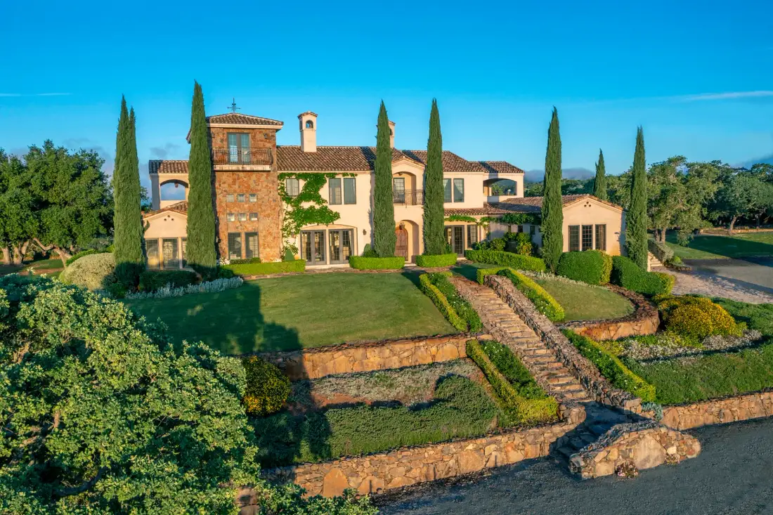 A large countryside house stands above a landscaped garden with stone terraces.