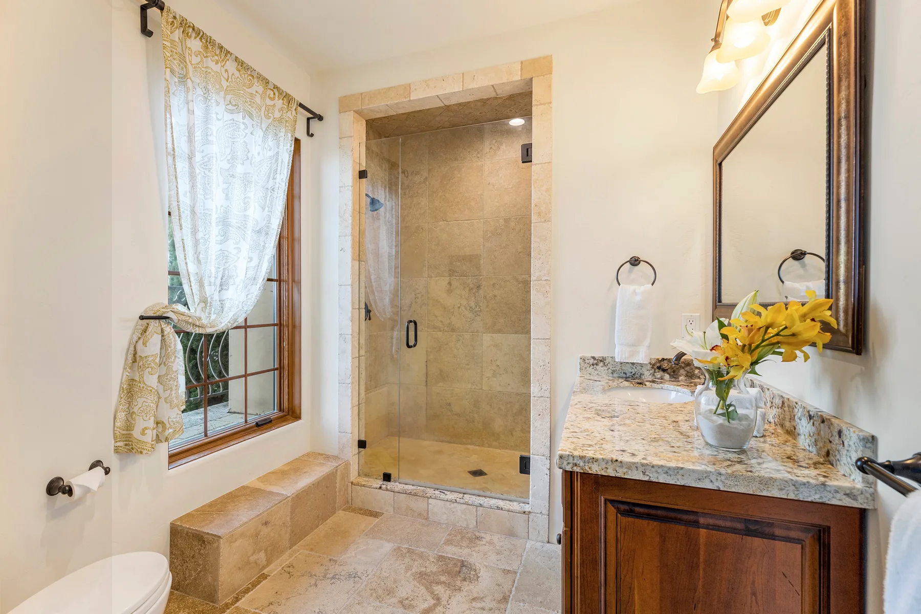 A bright bathroom with a glass shower, vanity, and curtained window.