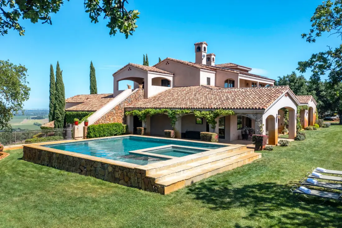 A Mediterranean-style house and swimming pool sit in a sunny landscaped yard.