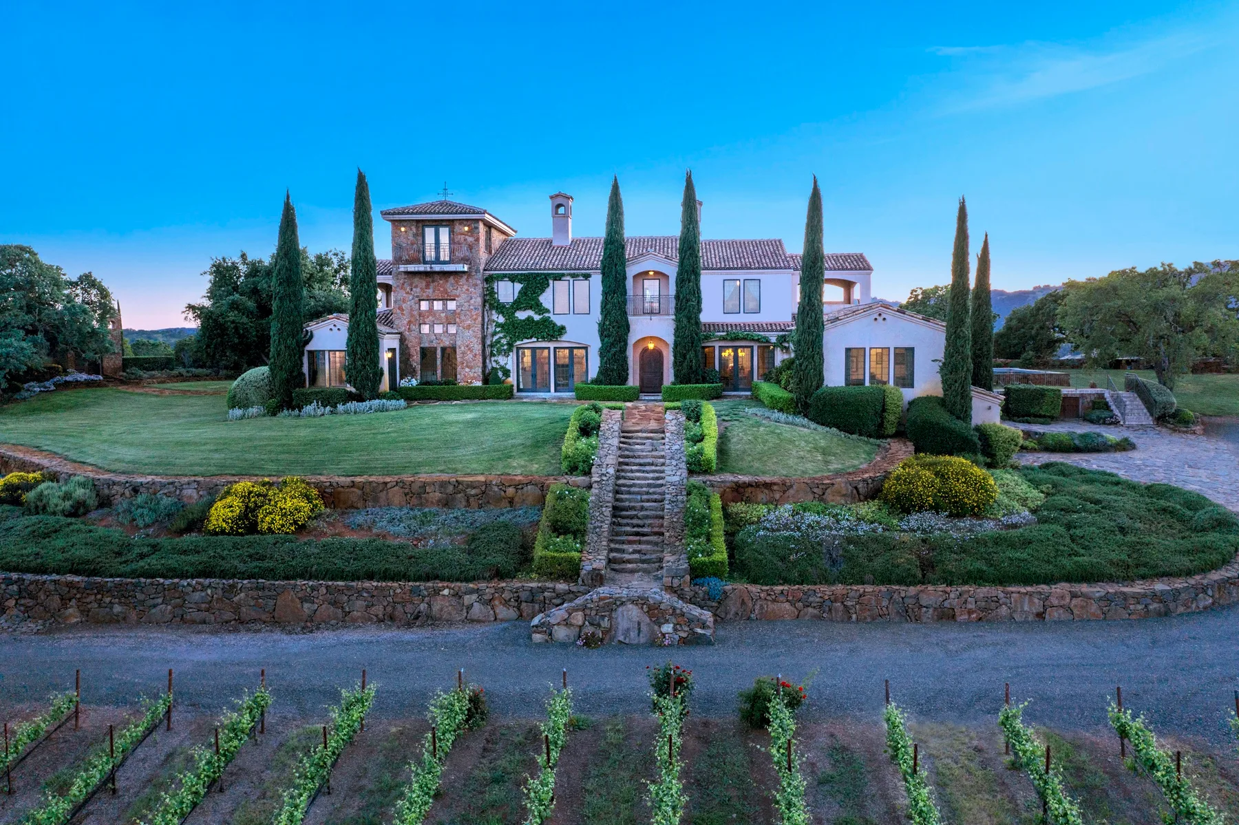 A large estate house stands beyond landscaped gardens and rows of vines.