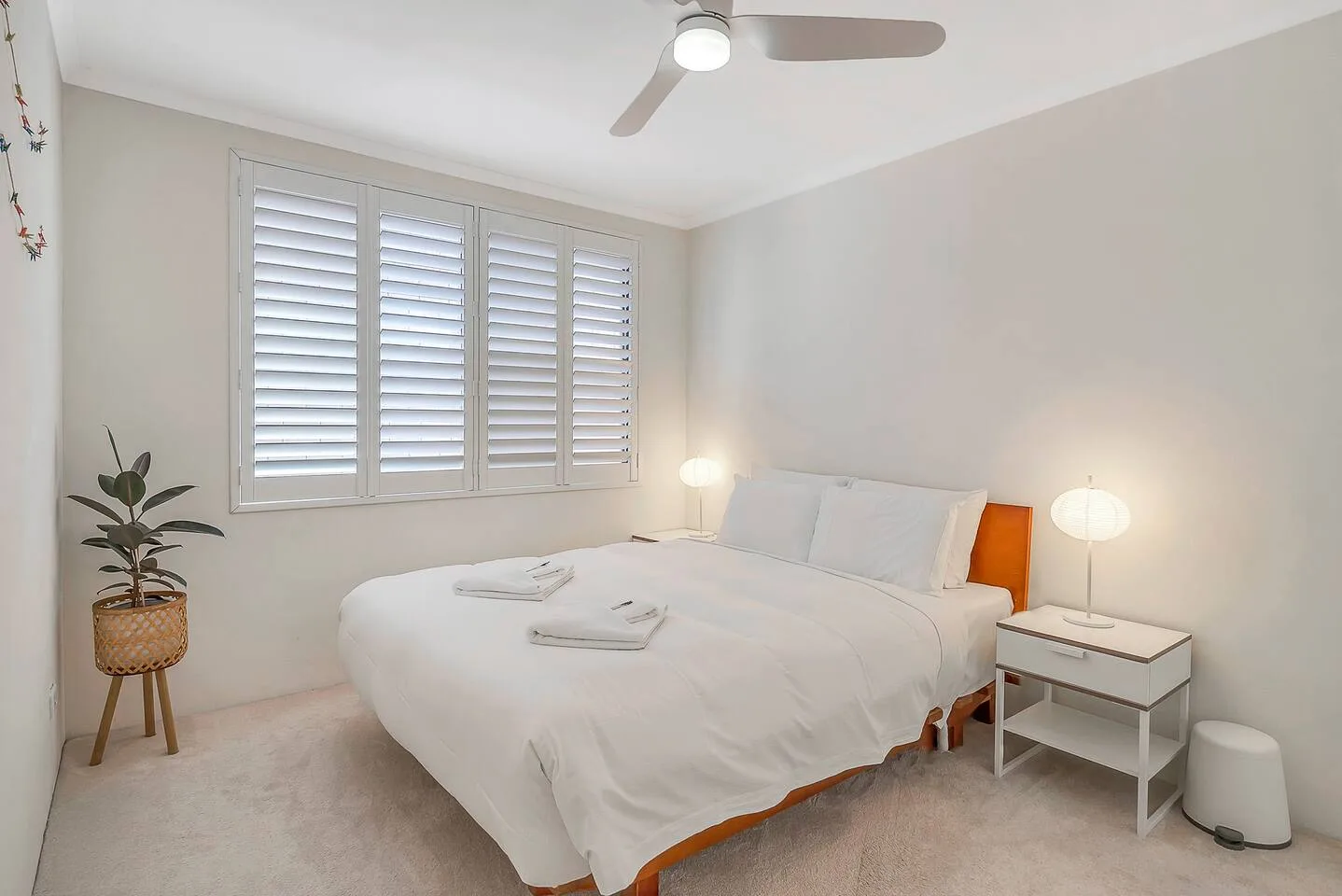 A bright bedroom with a neatly made bed, shuttered window, and ceiling fan.