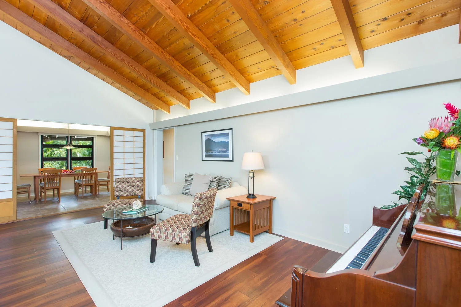 A bright living room with a seating area, piano, and exposed wooden ceiling.