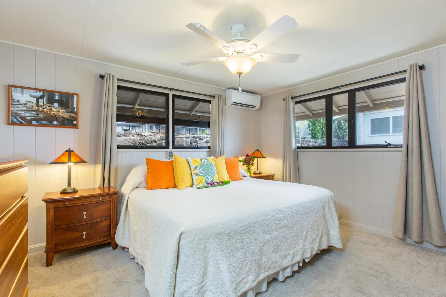 Un dormitorio con una cama, almohadas de colores y ventanas en dos paredes.