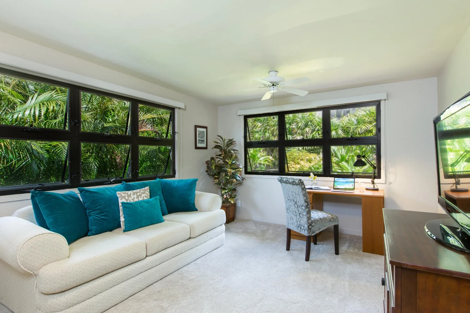 A bright sitting room with teal cushions and a desk beside large windows.