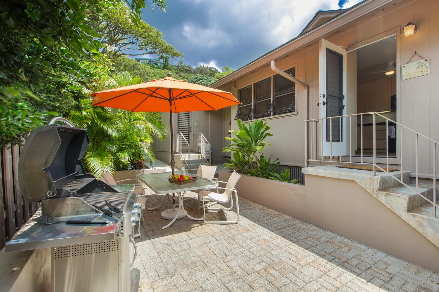 An outdoor patio with a table, orange umbrella, and barbecue beside a house.