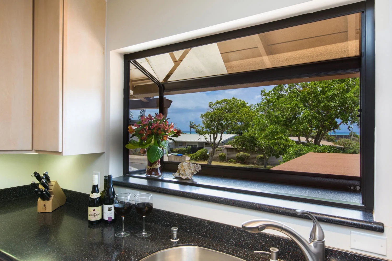 Un fregadero y una encimera de cocina están bajo una ventana amplia con vistas a árboles frondosos y tejados.
