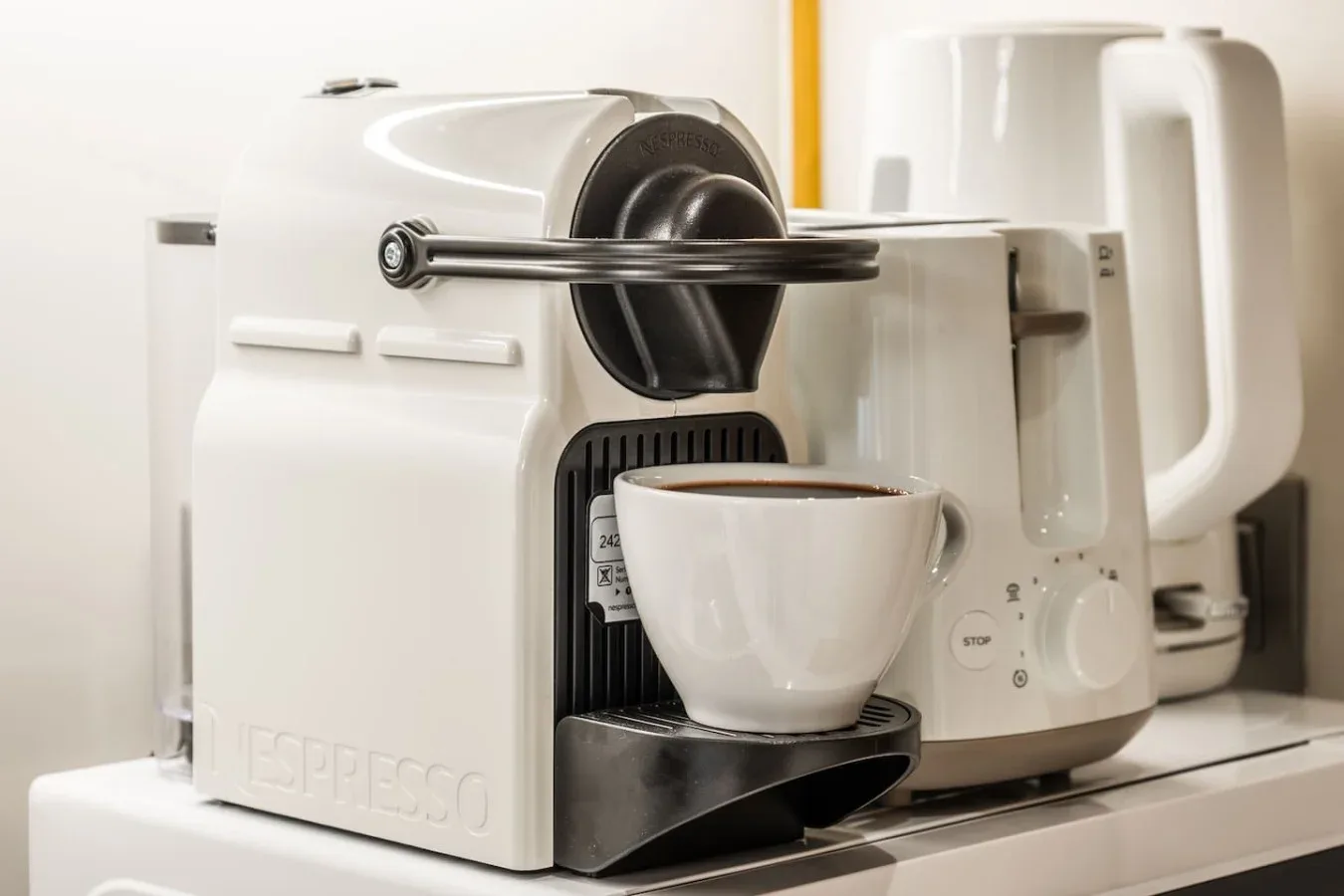 Una cafetera sobre la encimera sirve café en una taza blanca junto a un hervidor.