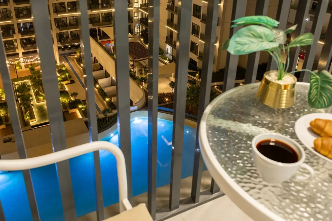 Una mesa de balcón con café y una planta da a una piscina iluminada de azul.