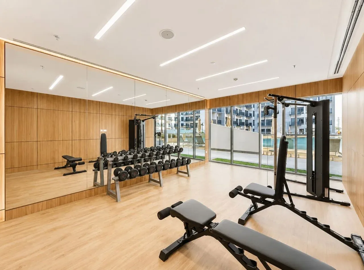 Sala de ejercicio con bancos y pesas junto a paredes con espejos y grandes ventanales.