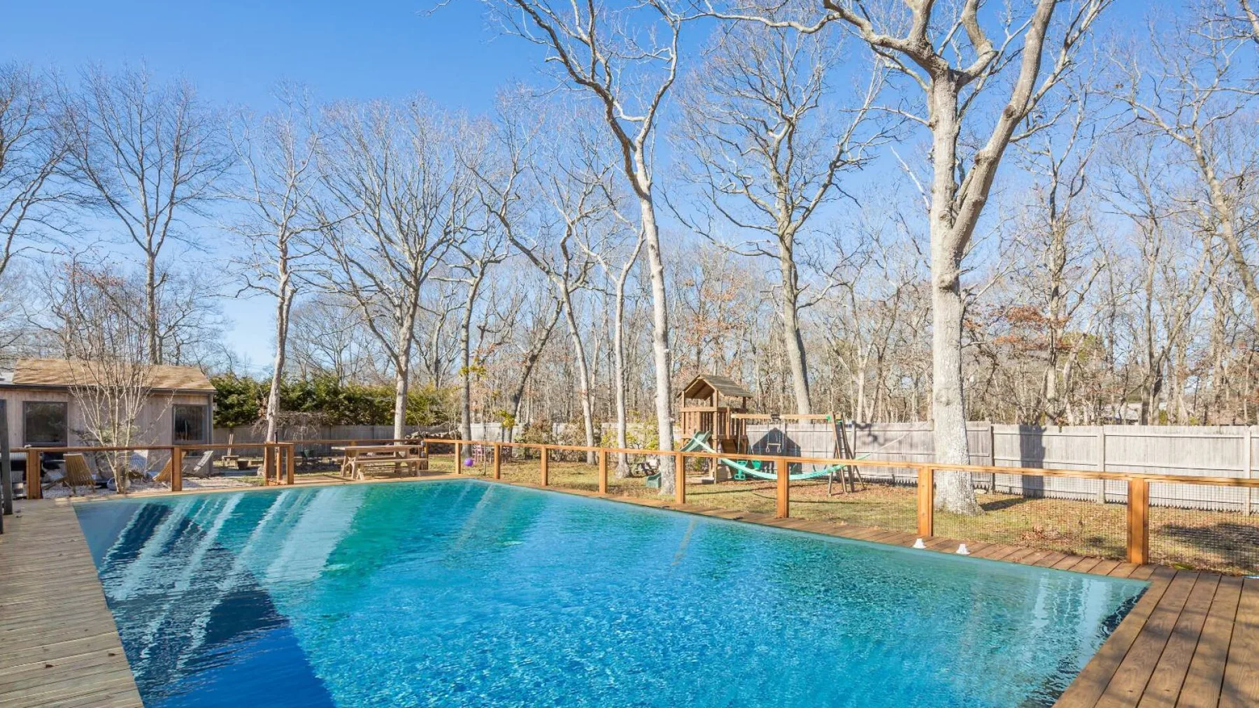 Una piscina está junto a una terraza de madera y árboles sin hojas.