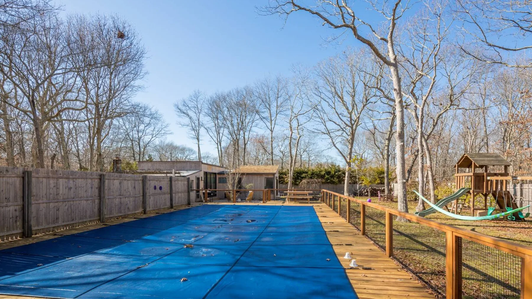 Zona de piscina exterior cercada, con una terraza de madera y árboles sin hojas.