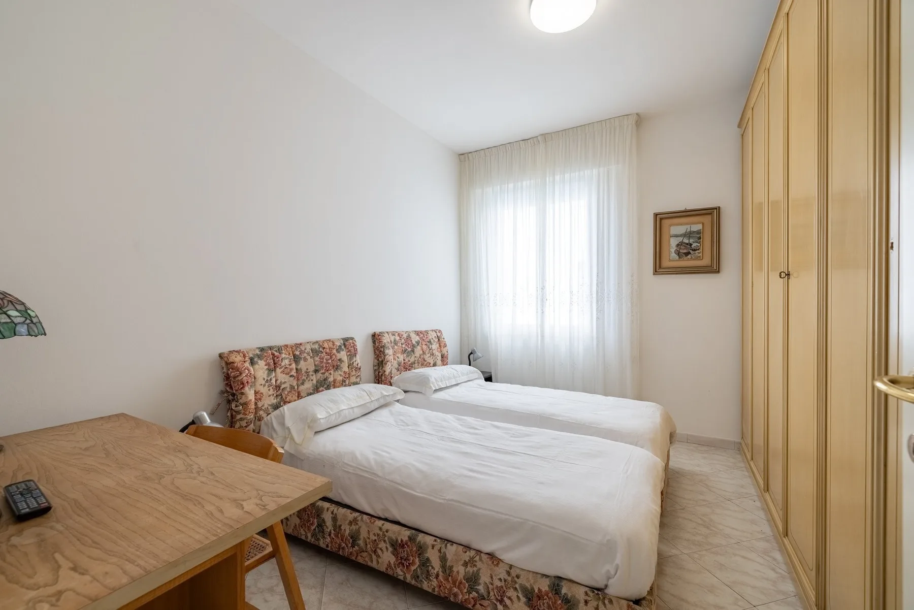 A bedroom with two beds, a wooden desk, and a tall wardrobe.