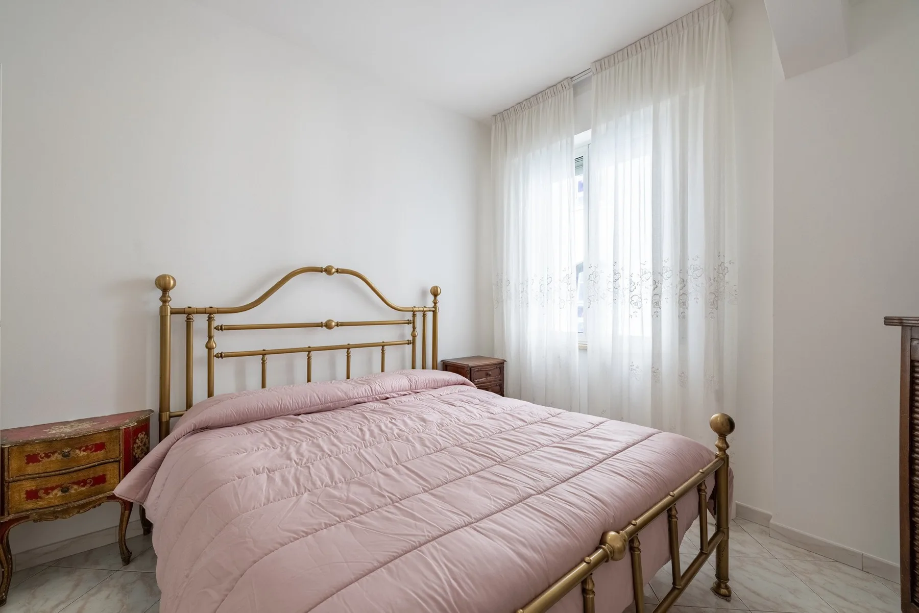 A bedroom with a brass bed dressed in pink bedding beside a curtained window.
