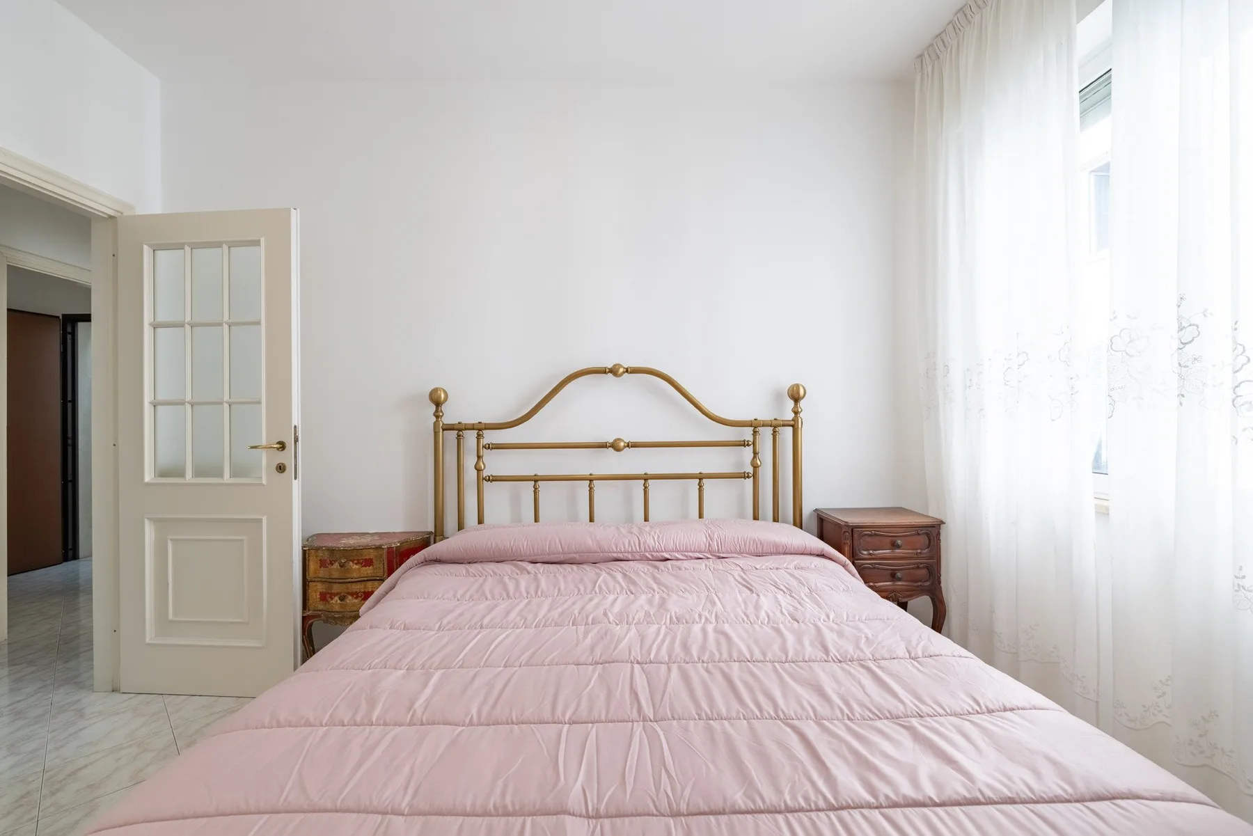 A bright bedroom with a pink bed, brass headboard, and tall curtained window.