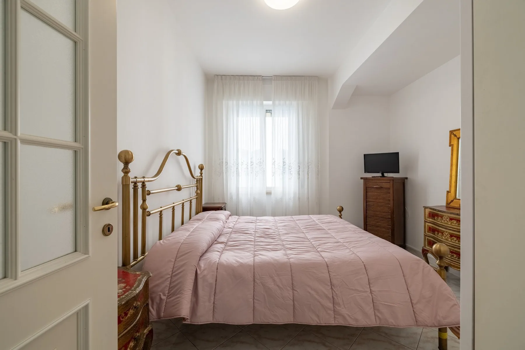 A bedroom with a brass bed, pink bedding, and a television by the window.