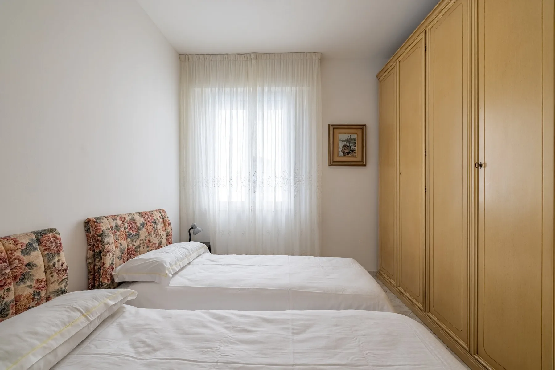 A bedroom with two beds, a large wooden wardrobe, and a curtained window.