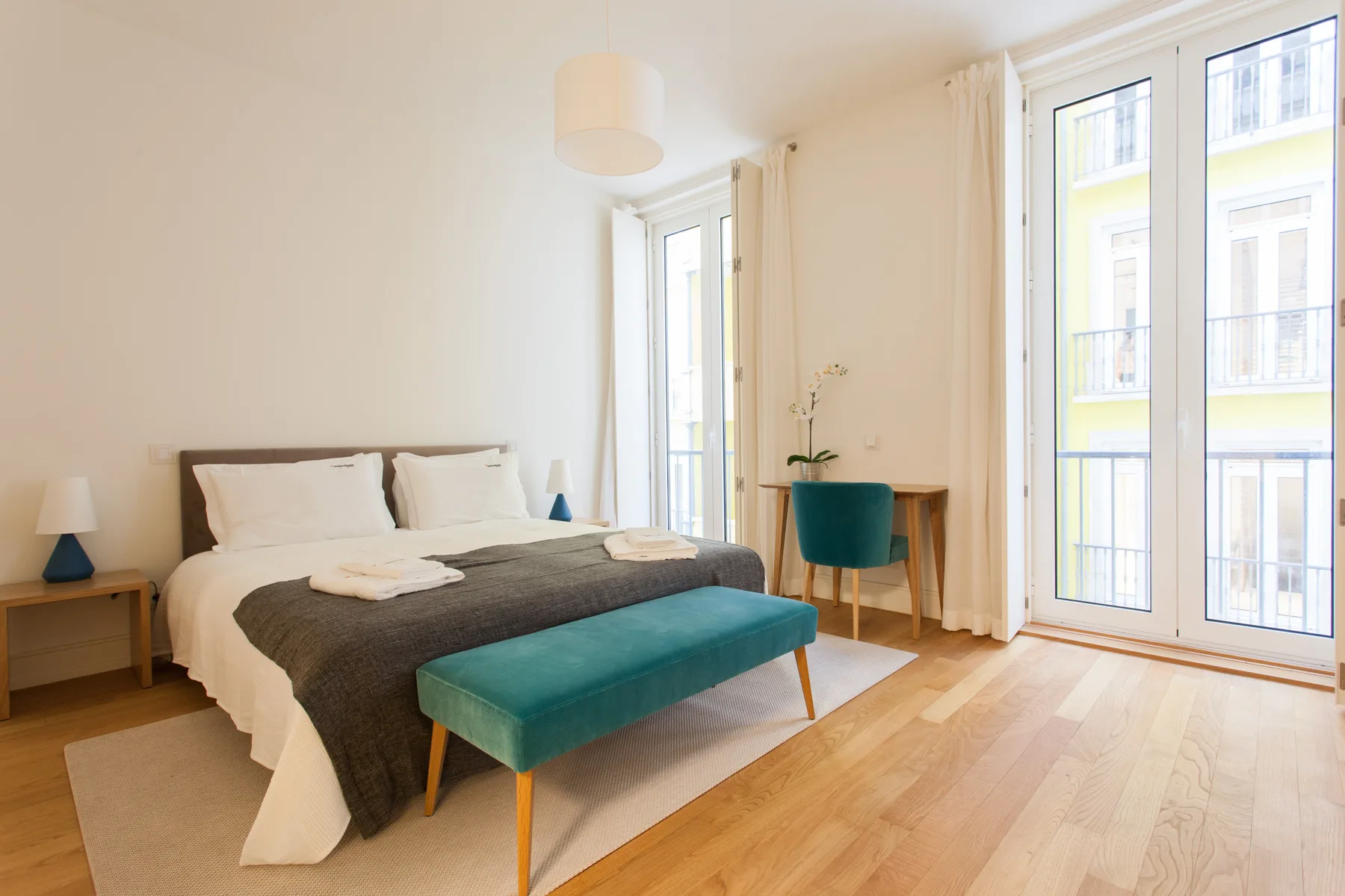 A bright bedroom with a neatly made bed, a teal bench, and tall windows.