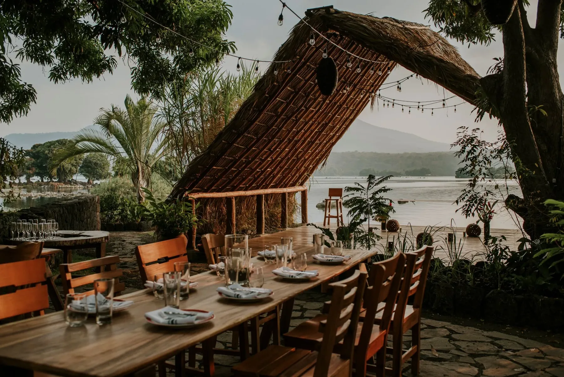 Outdoor dining table set under a thatched roof beside the water.