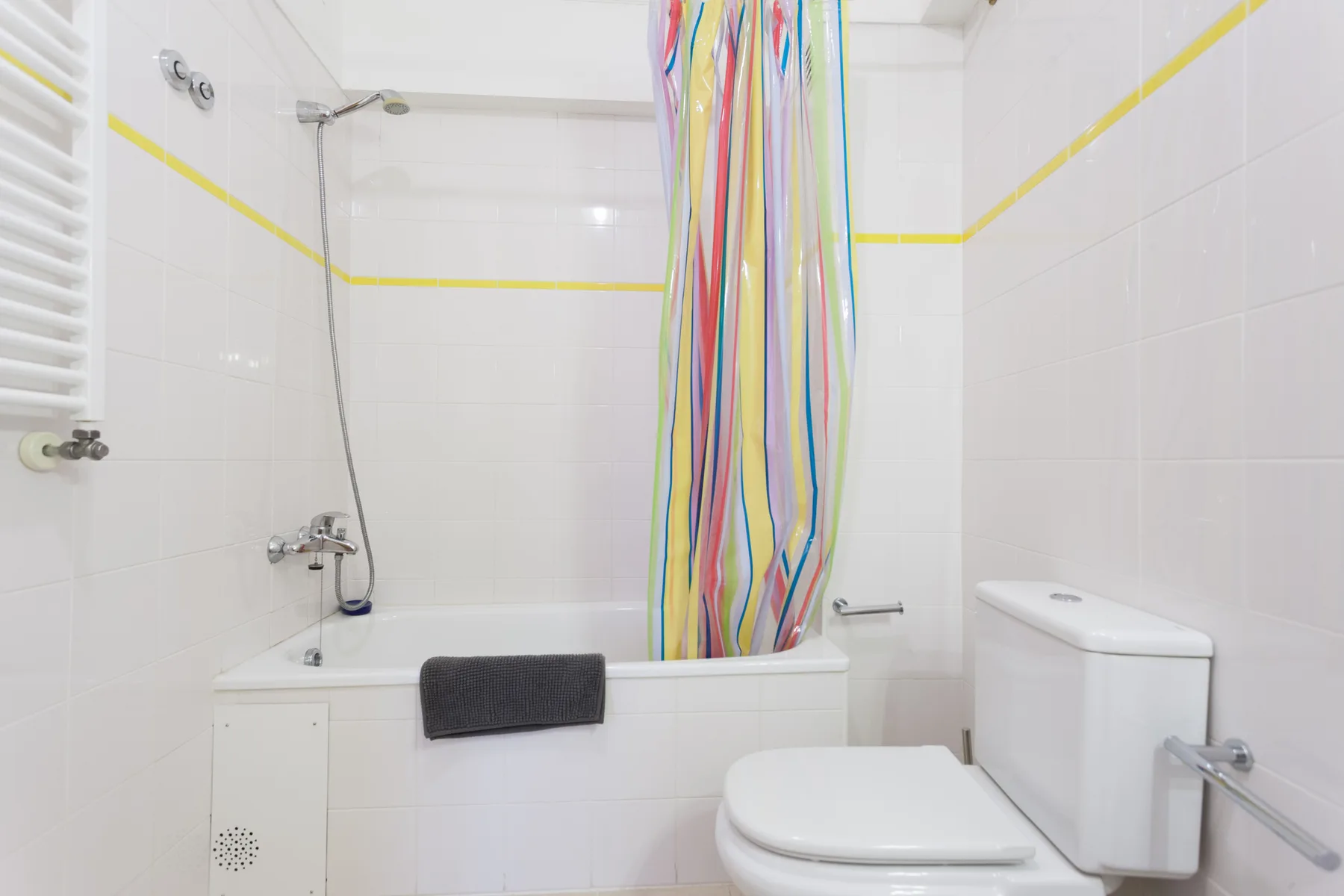 Un baño de azulejos blancos con bañera, cortina de ducha colorida e inodoro.