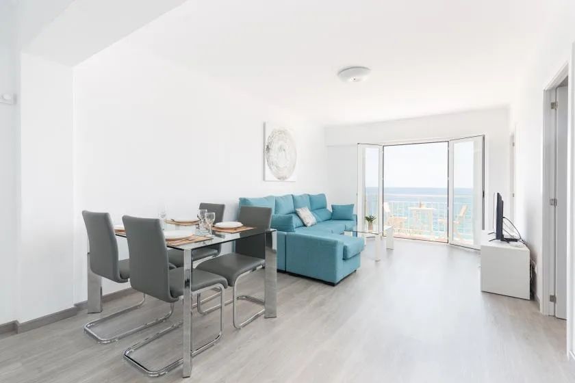 A bright living and dining area with teal seating and large windows overlooking the water.