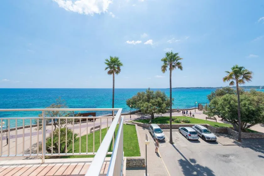A seaside view from a balcony overlooks the blue water and nearby parking area.