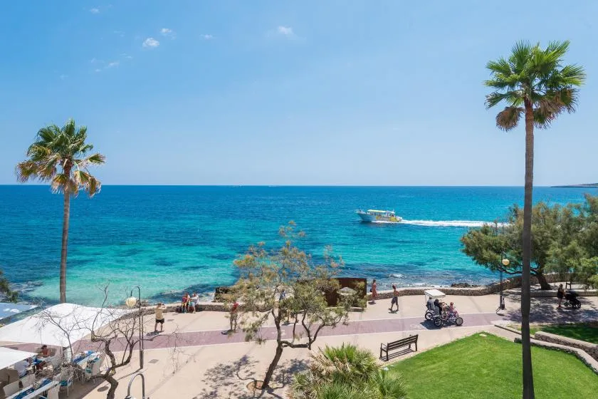 A seaside promenade overlooks turquoise water, with palm trees and a boat offshore.