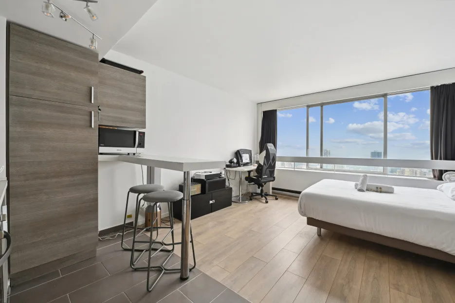 Un dormitorio luminoso con una cama, un escritorio y grandes ventanas con vistas a la ciudad.