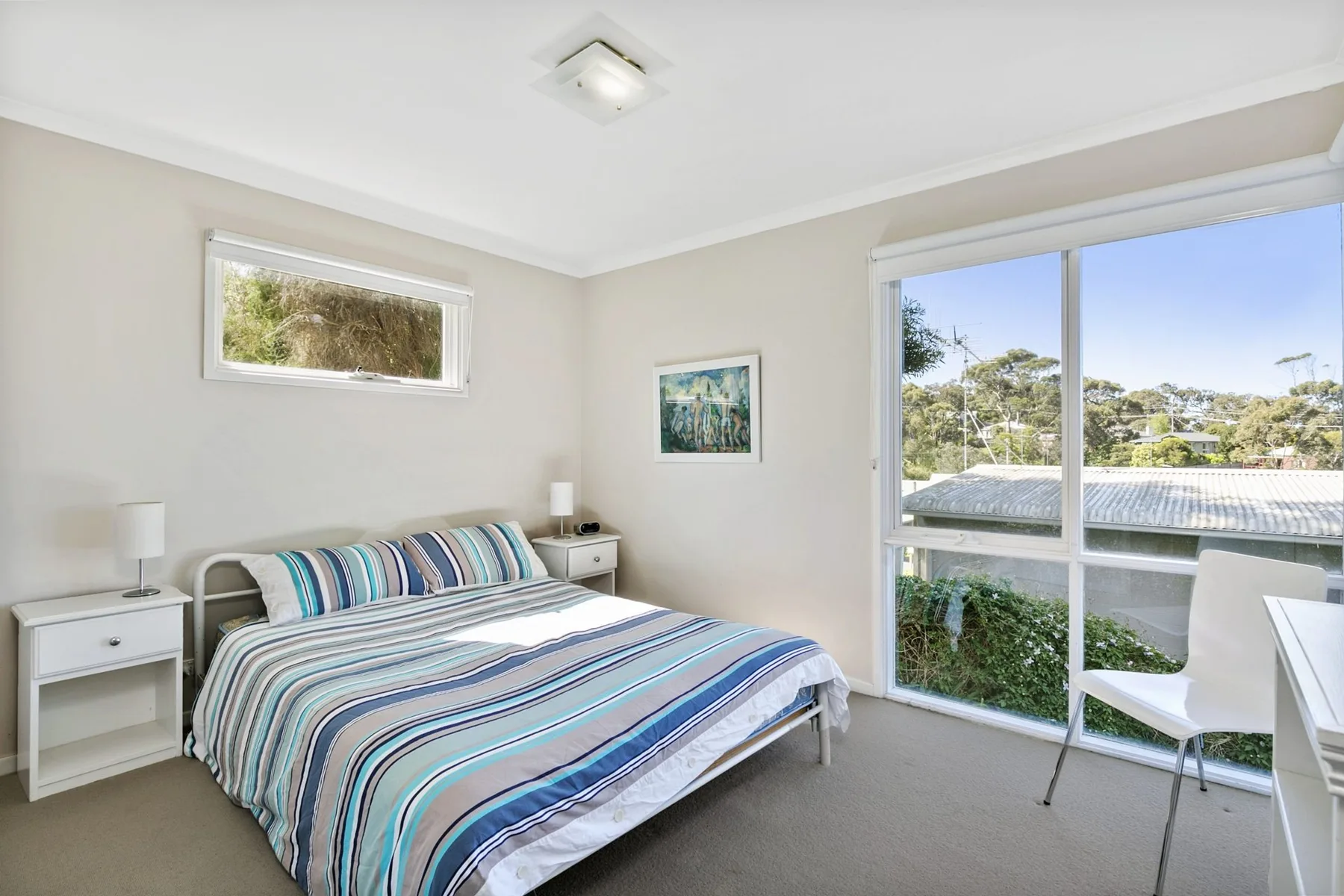 A bright bedroom with striped bedding and large windows overlooking greenery.