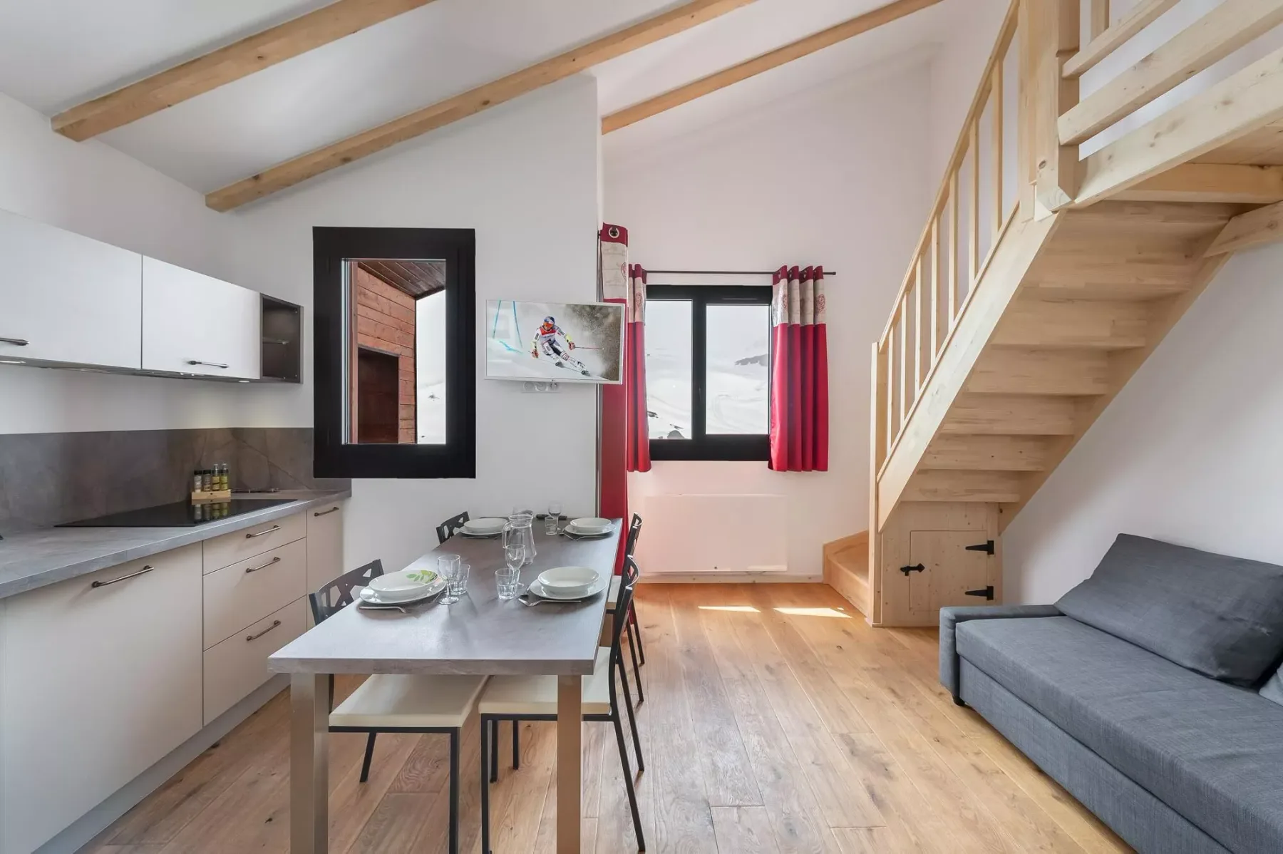 A bright open-plan room has a kitchen, dining table, and staircase beside a gray sofa.