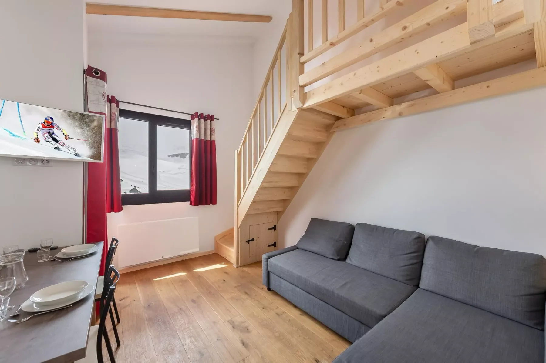 A living area with gray seating beneath a wooden loft staircase.