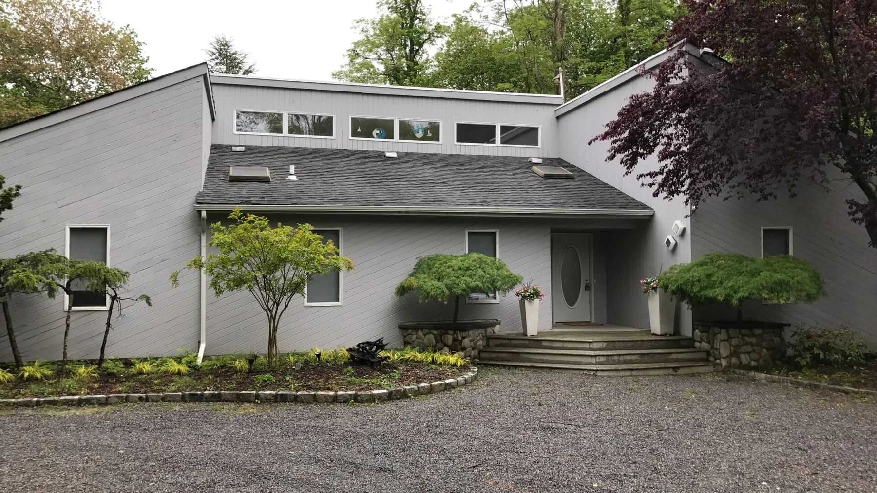 A gray house exterior with a gravel driveway and landscaped shrubs.