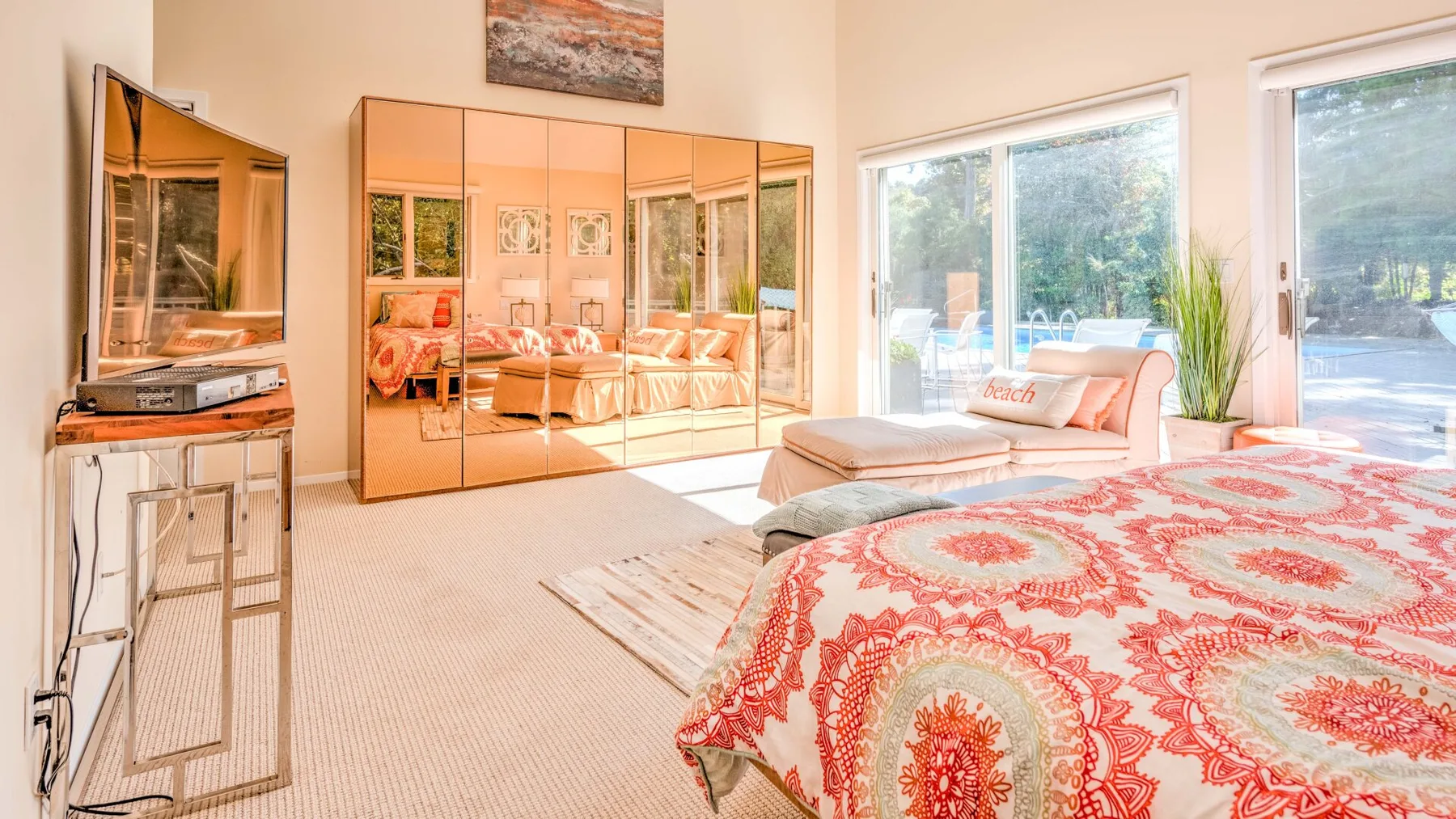A bright bedroom with a patterned bedspread, mirrored wardrobe, and large windows.