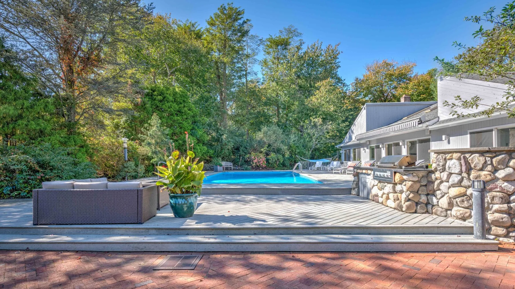 Una piscina al aire libre junto a una terraza, con asientos y árboles alrededor.
