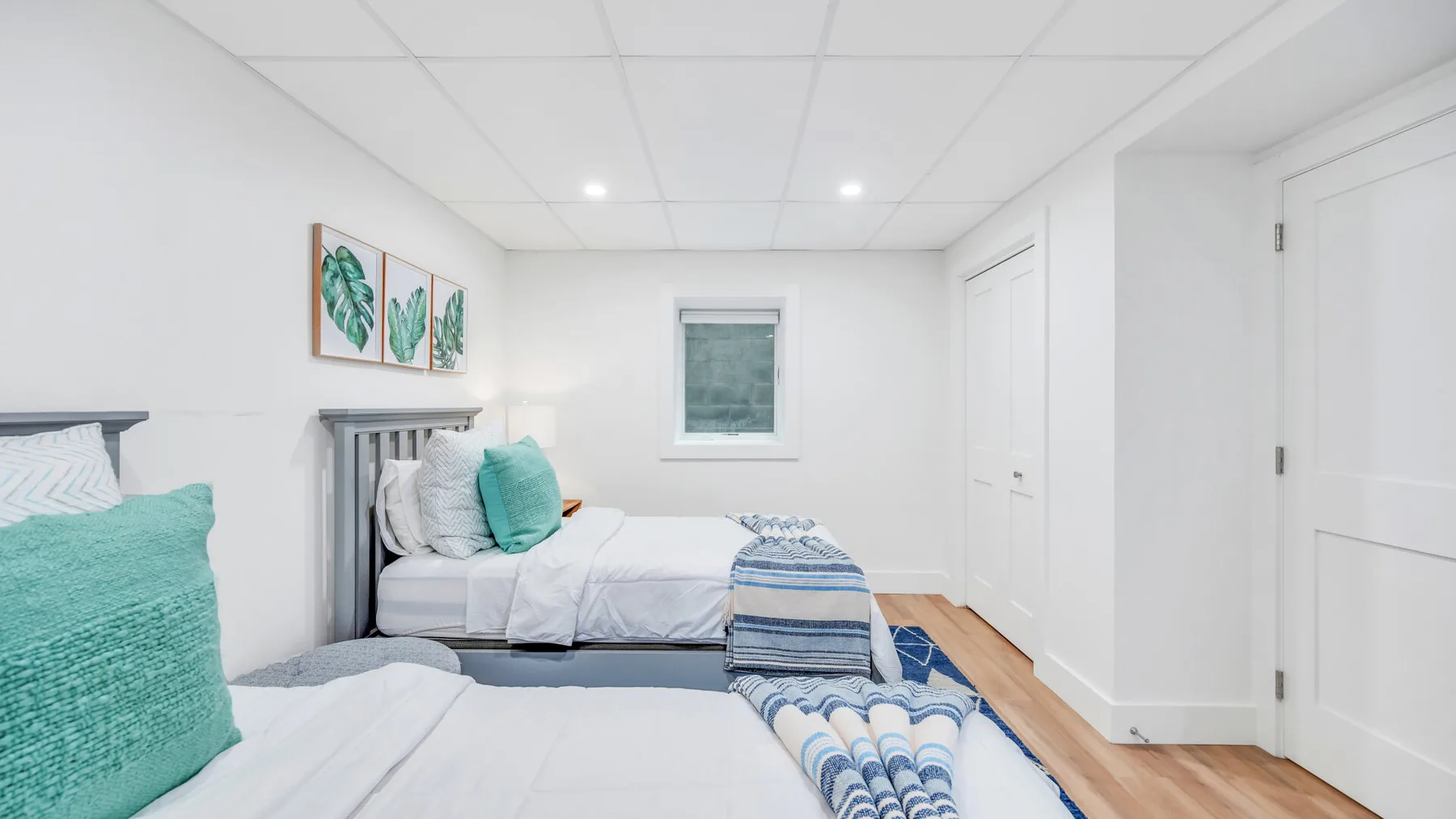 A bright bedroom with two beds, teal pillows, and blue-and-white bedding.