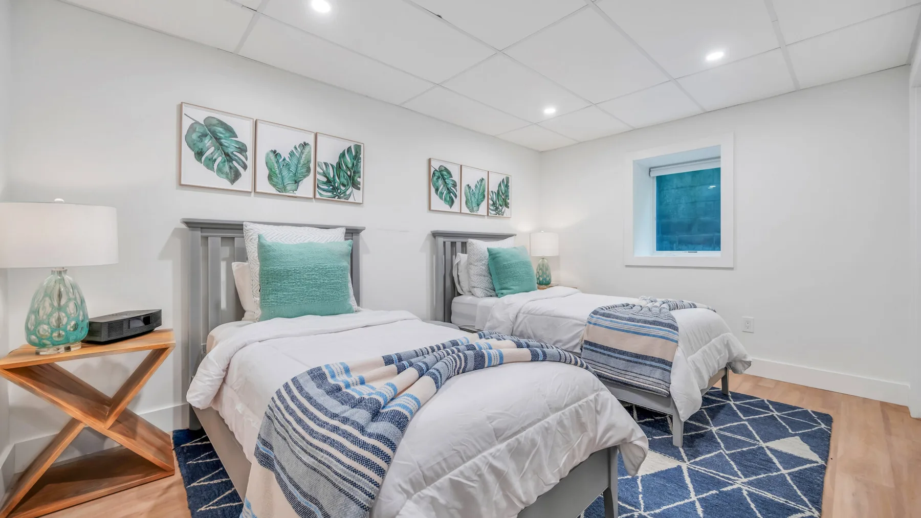 A bright bedroom with two beds, teal accents, and botanical wall prints.
