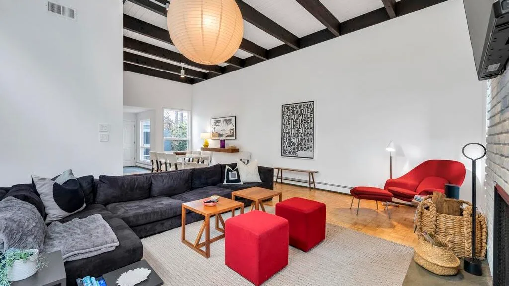 A bright living room with sectional seating, red ottomans, and exposed ceiling beams.