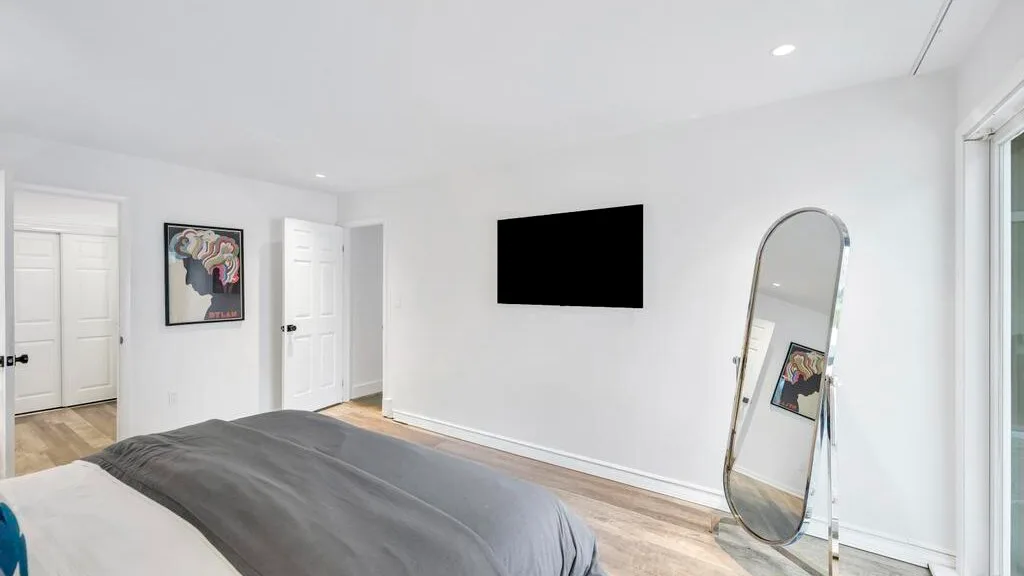 A bright bedroom with a bed, wall-mounted television, and full-length mirror.