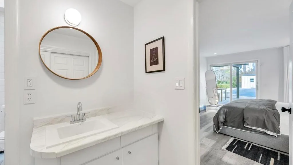 A bathroom vanity with a round mirror overlooks a bedroom with a bed.