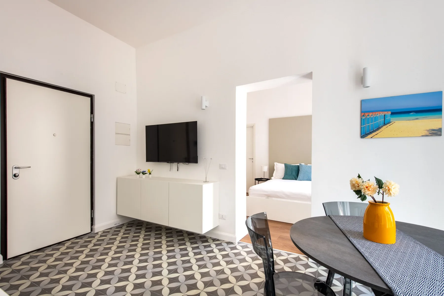 Bright dining area with a round table, wall-mounted television, and view into a bedroom.