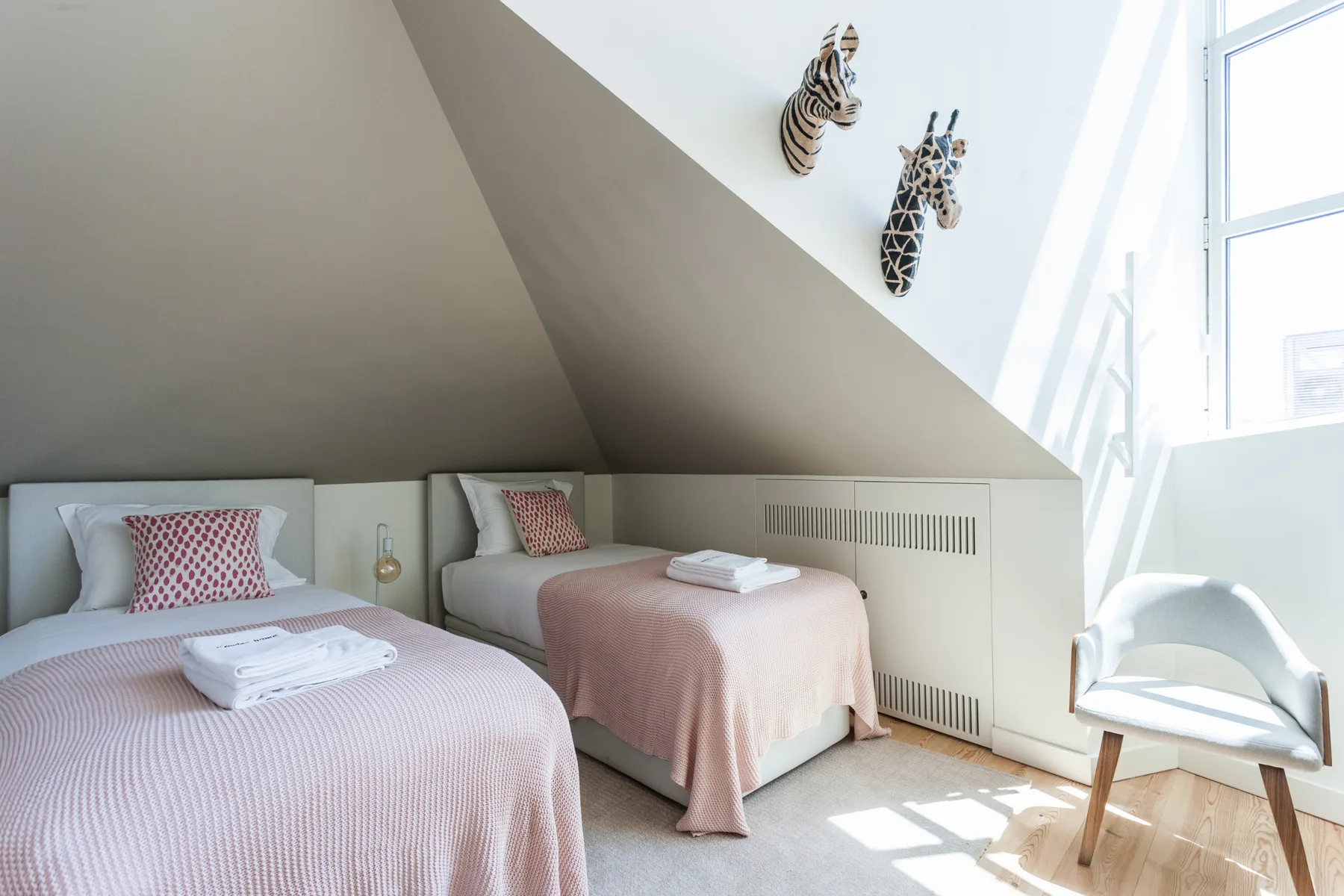 Un dormitorio luminoso con dos camas y una silla junto a una ventana grande.