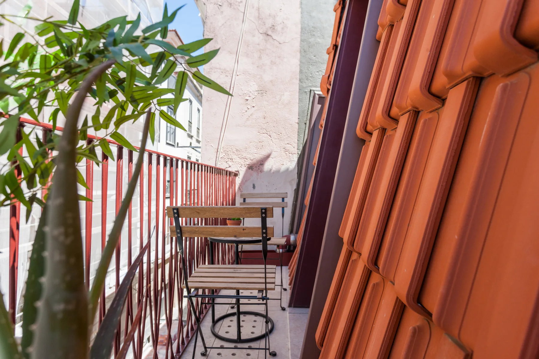 A narrow balcony with a small table and chairs beside a leafy potted plant.