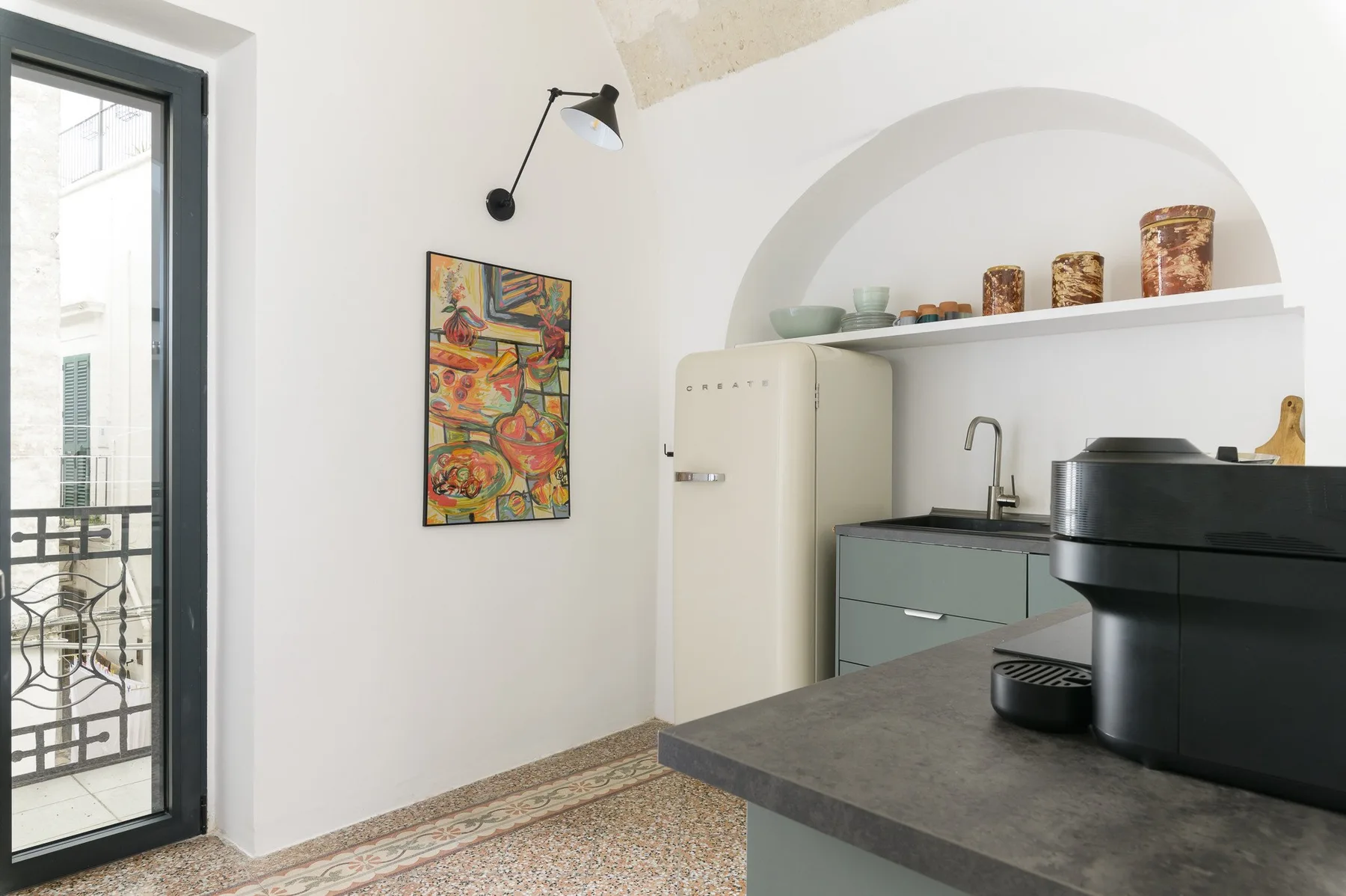 Una cocina luminosa con encimera oscura, refrigerador y nicho arqueado en la pared.
