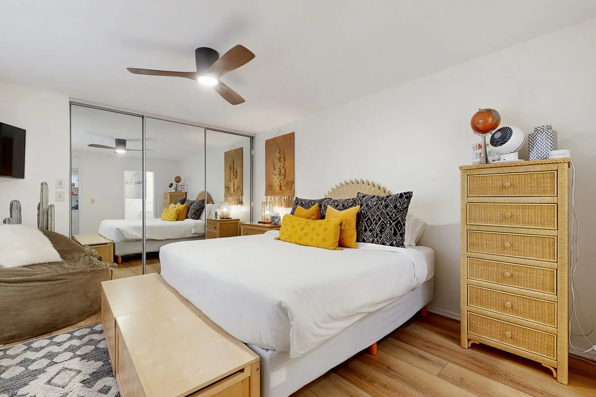 A bright bedroom with a bed, mirrored wardrobe, and ceiling fan.